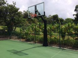 Basketball hoop on green court with black pole. Trees and fence in the background. Cloudy sky.