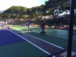 Two workers resurfacing a tennis court with green and purple paint, sunny day.