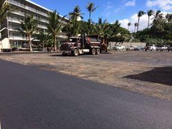 Asphalt paving a parking lot, dump truck present near a building and palm trees.