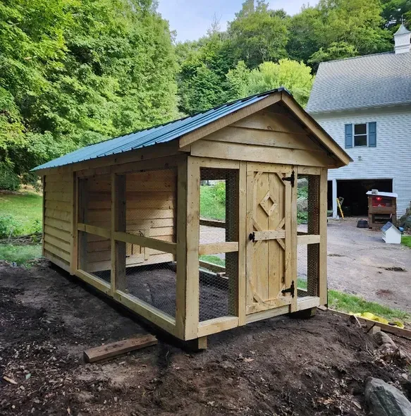 A small wooden shed with a blue roof is sitting in the dirt in front of a house.