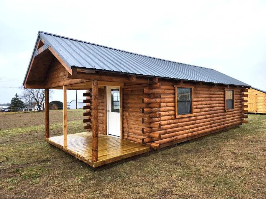 A small log cabin with a porch and a metal roof is sitting in the middle of a field.