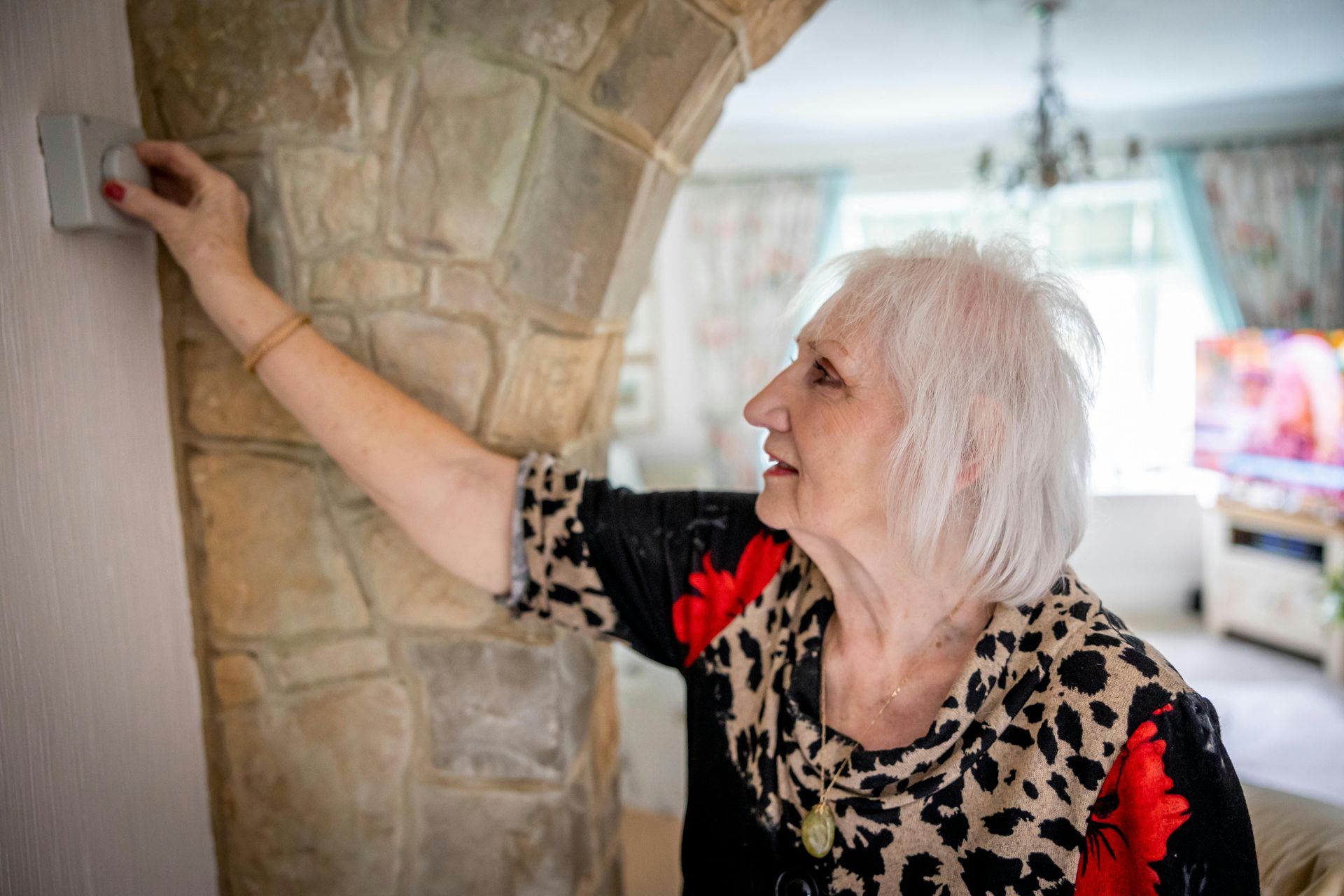 An elderly woman is adjusting a thermostat in a living room.