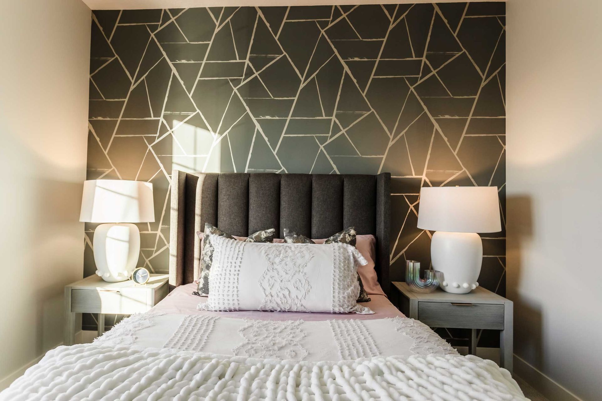 A bedroom with a bed , nightstands , lamps and a geometric wallpaper on the wall.