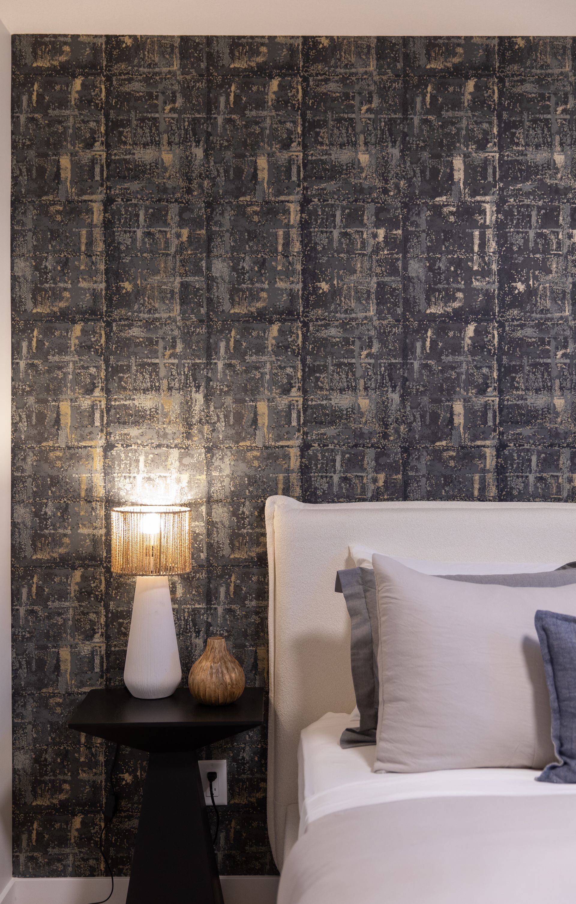 A bedroom with a bed , nightstand , lamp and wallpaper.