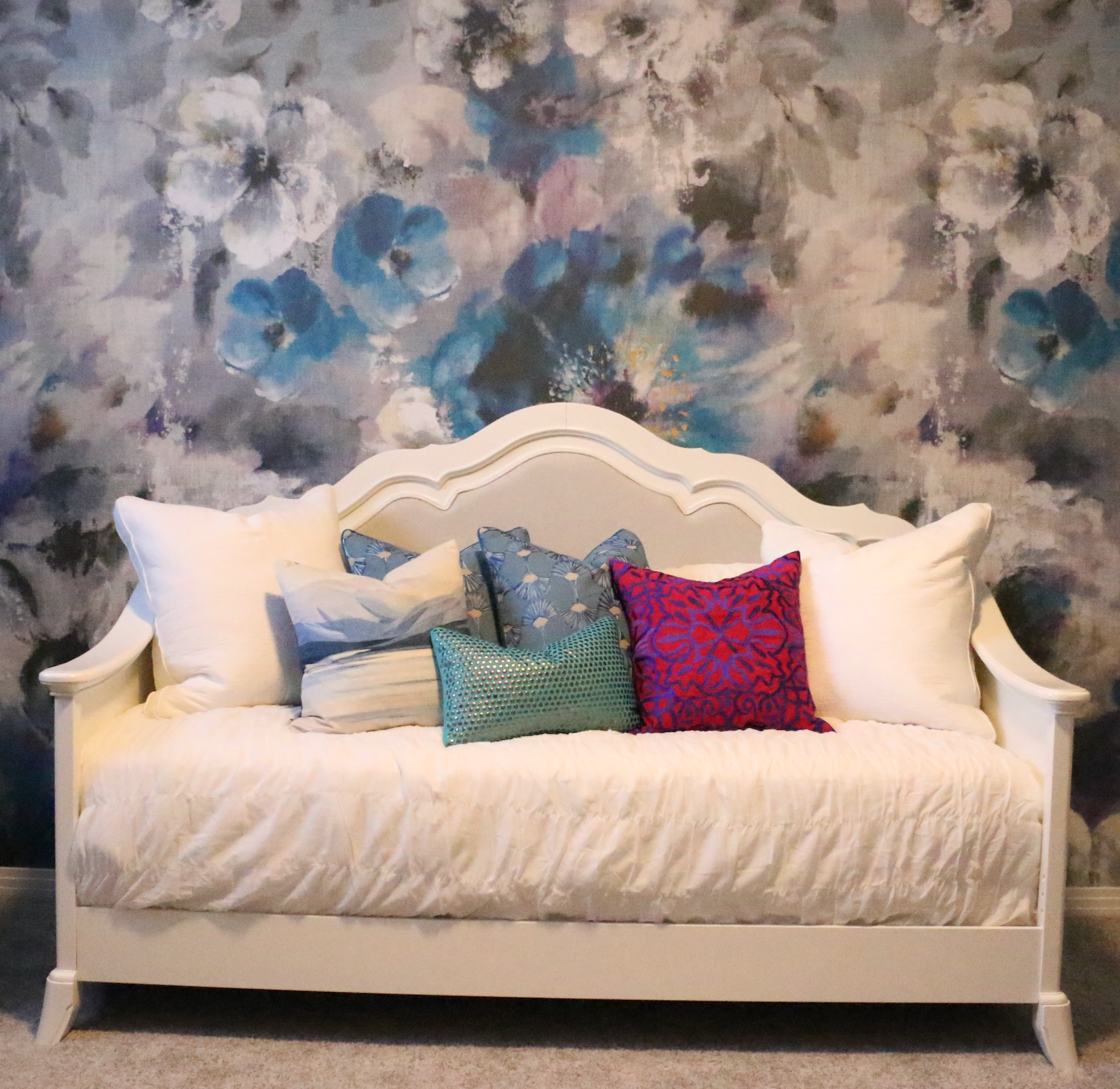 A white day bed with pillows on it in front of a floral wallpaper.