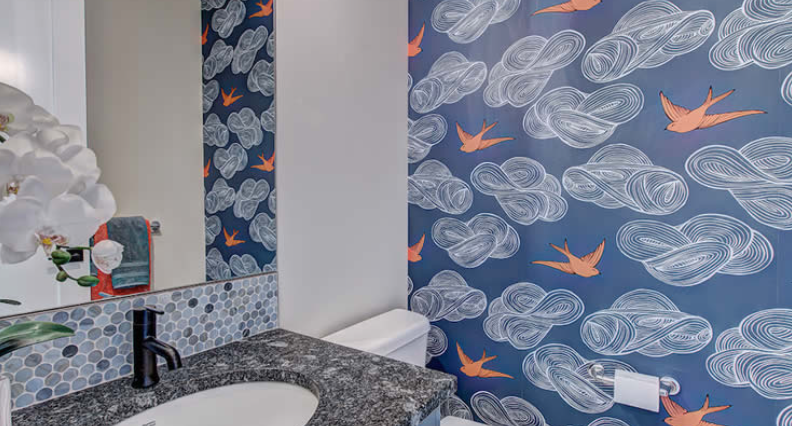 A bathroom with a sink , toilet , mirror and cloud and bird wallpaper.