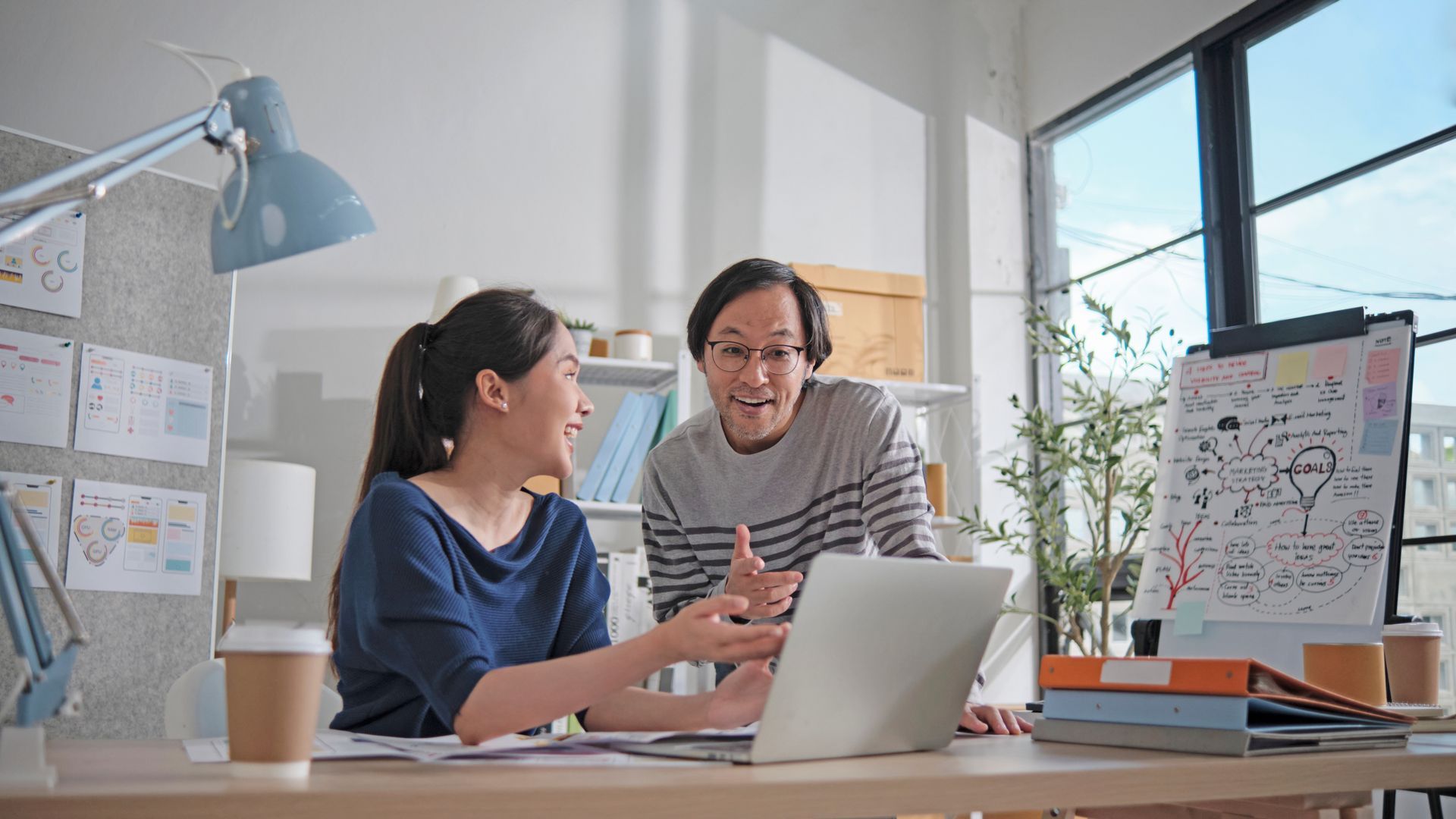 Two coworkers collaborating at a desk, laptop open, with sketches and notes; bright office setting.