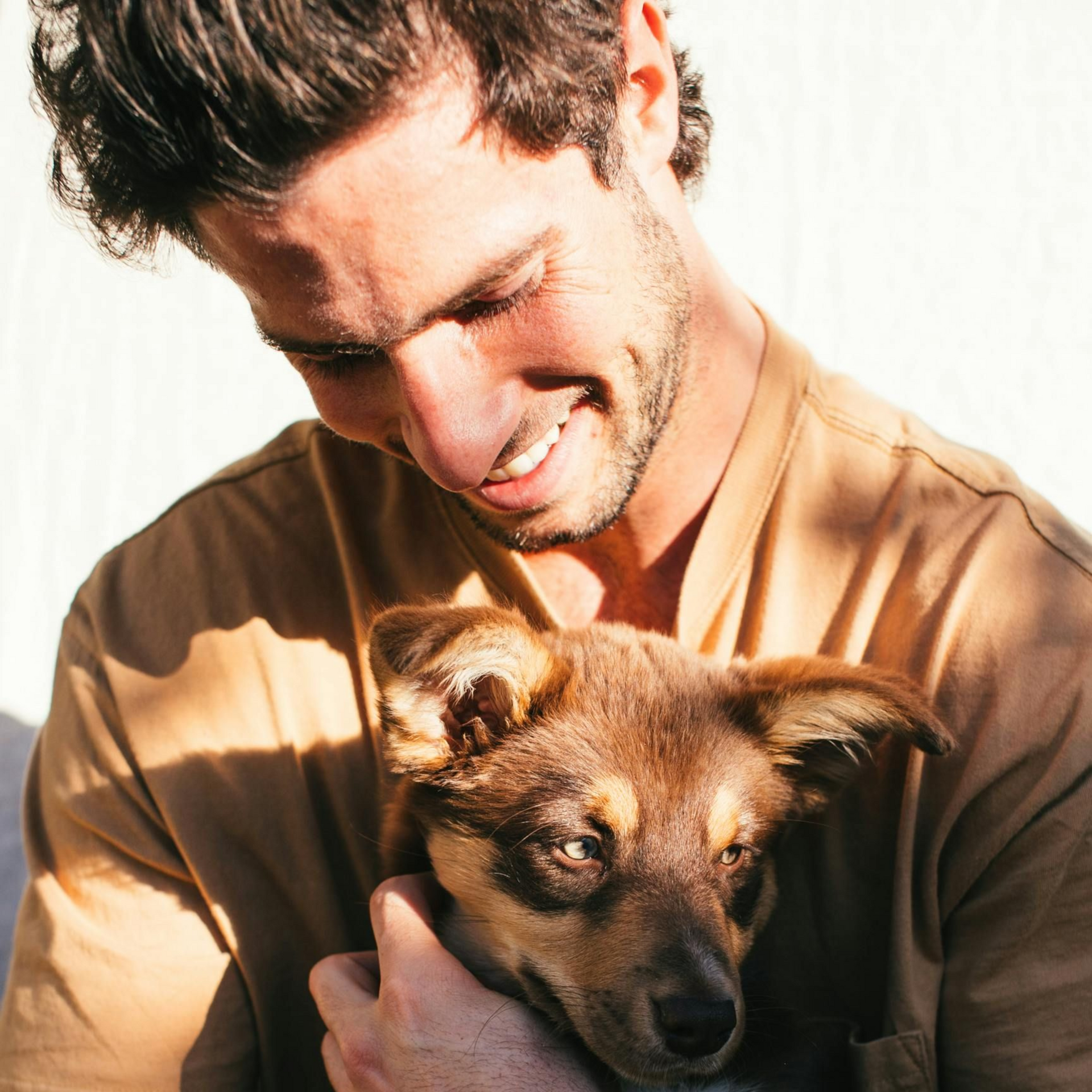 Man smiling while holding a brown puppy. Both looking down, natural light, beige shirt.