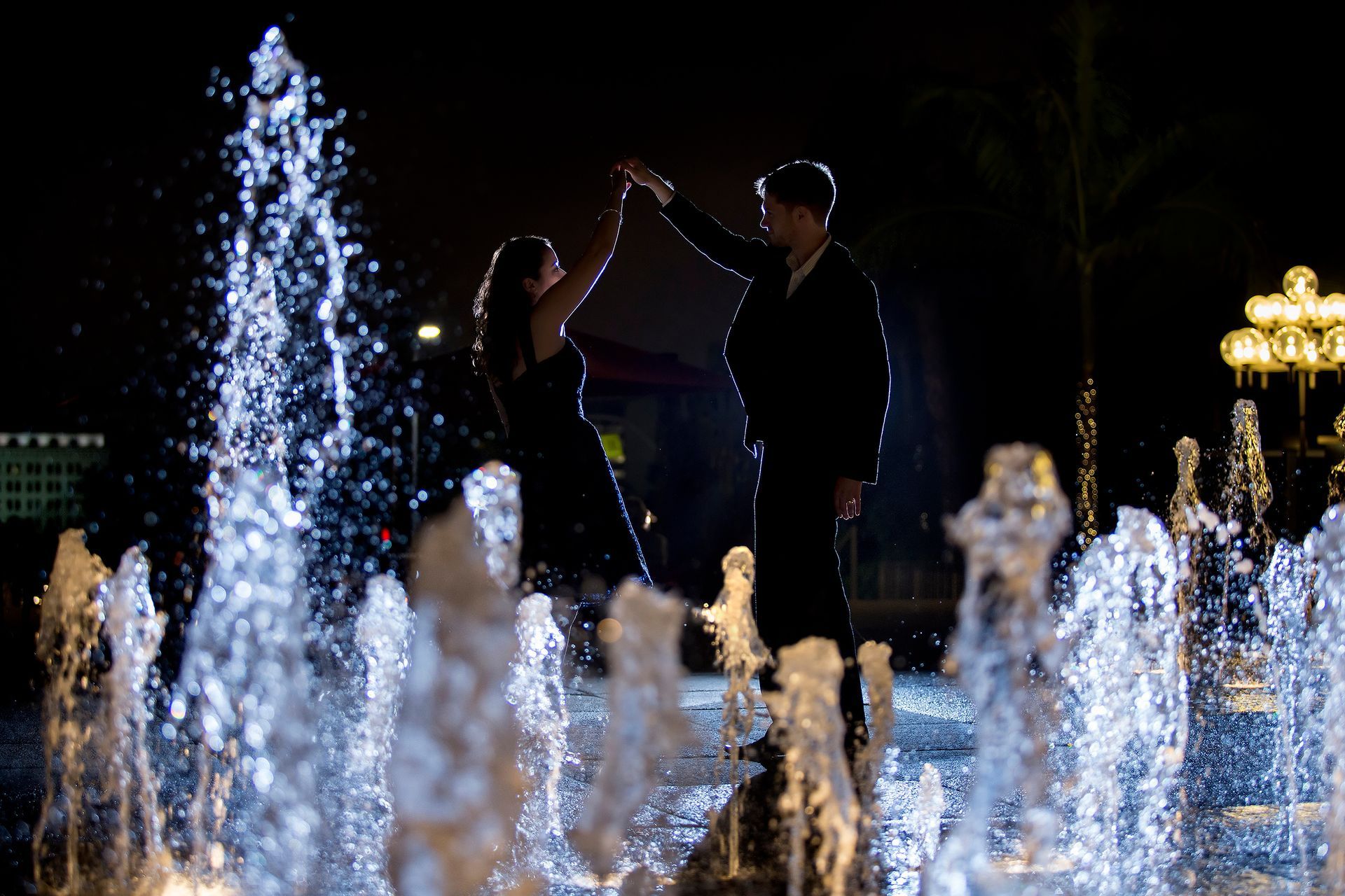 A man and a woman are dancing in a fountain at night.