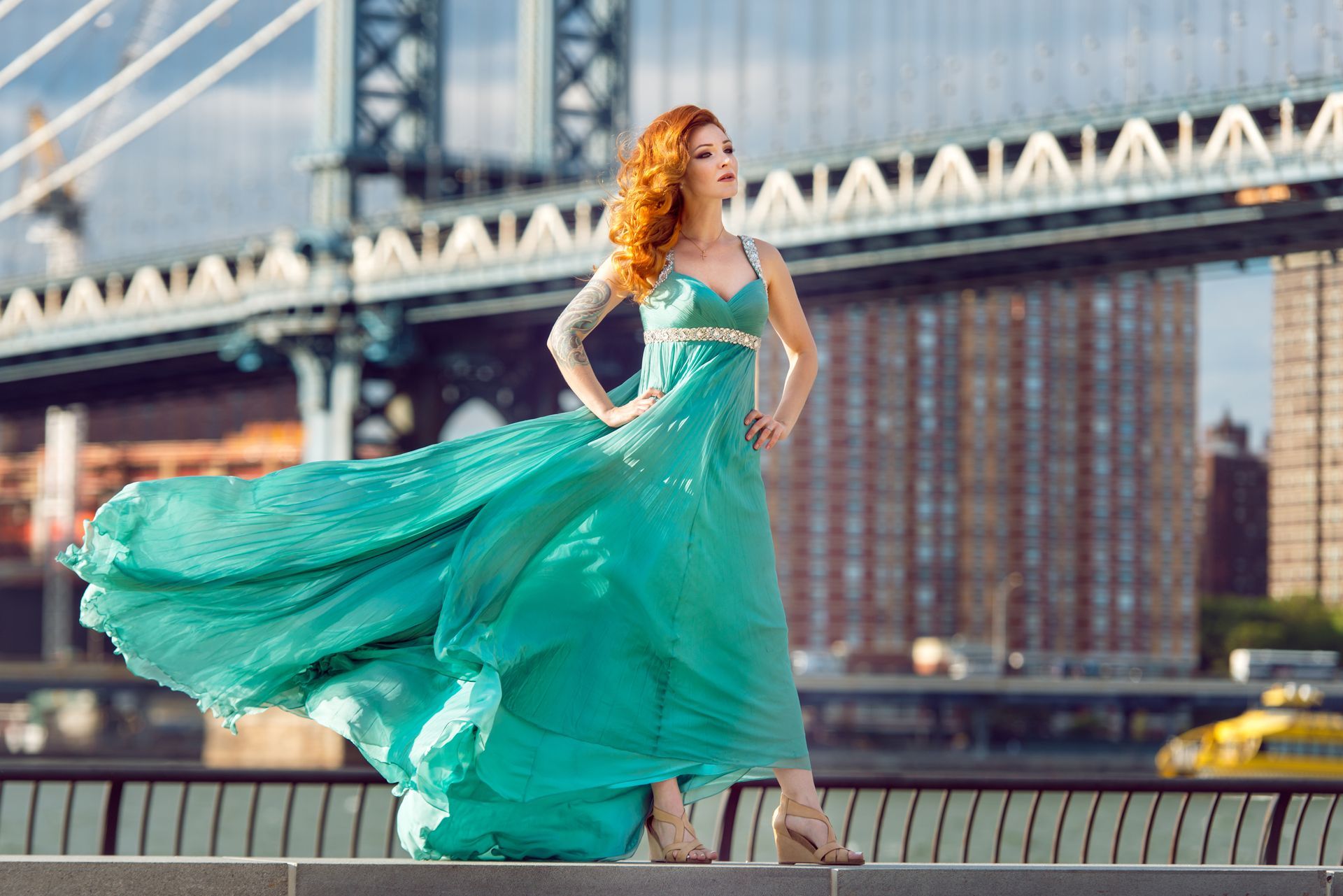 Woman in teal dress with flowing skirt, standing near bridge, New York City skyline in background.