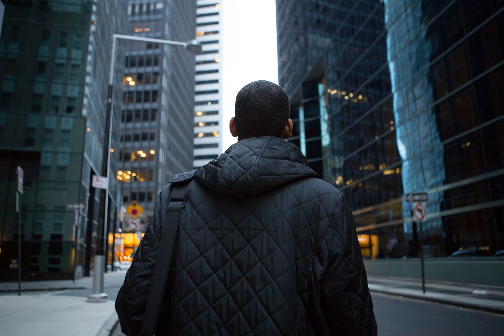 Man walking down city street, wearing black jacket, carrying a bag. Buildings line the street.