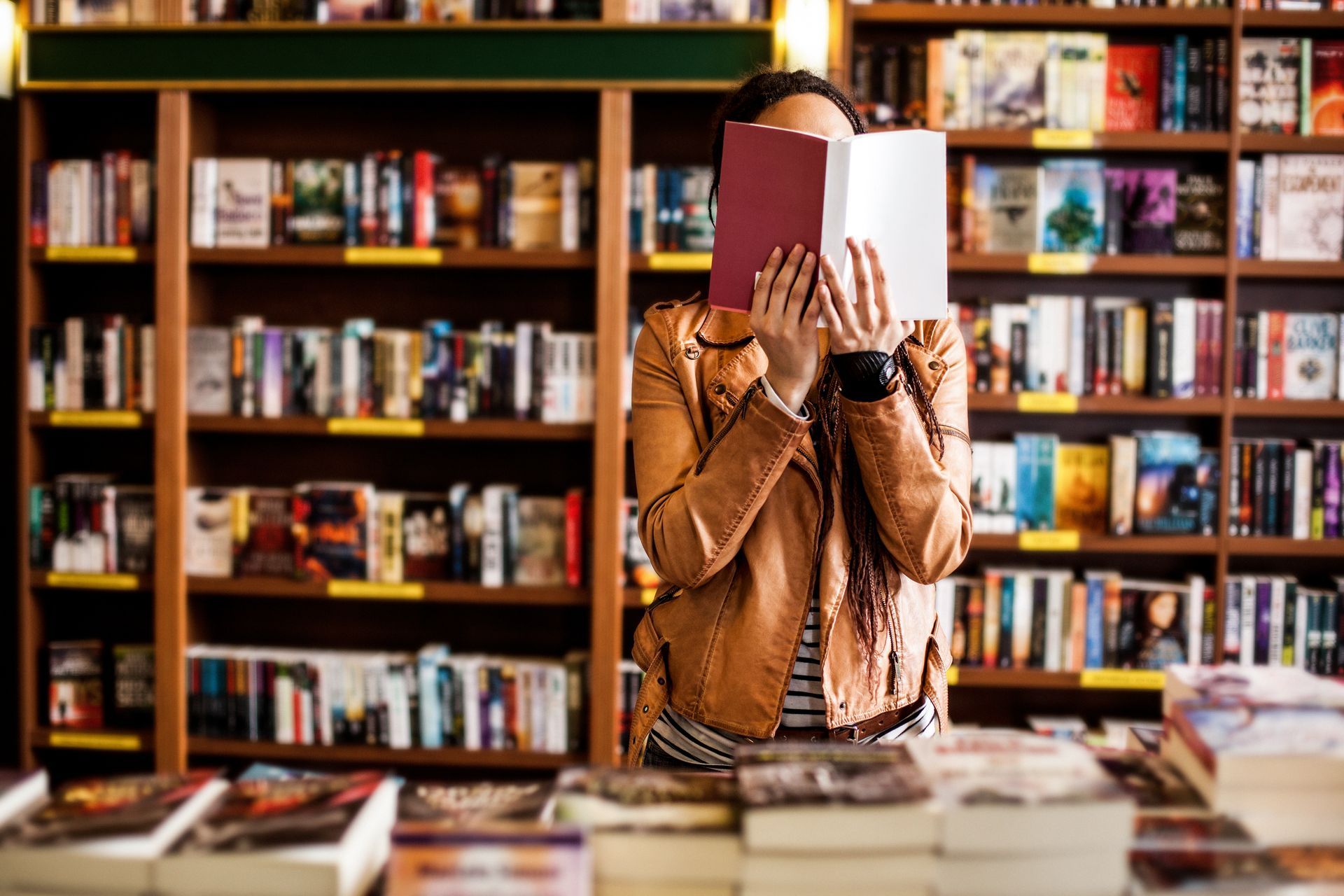 A woman is covering her face with a book in a bookstore.