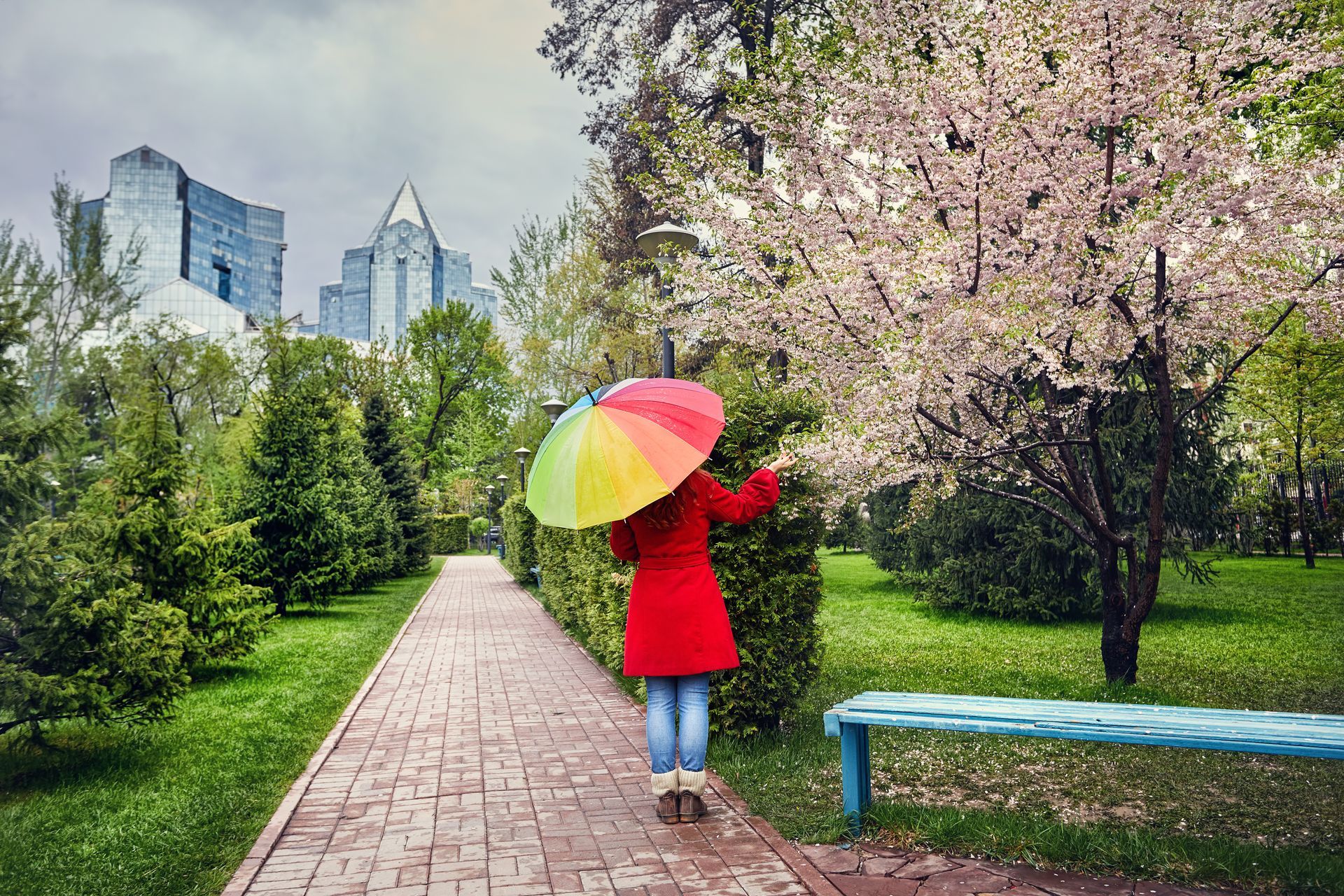 A person in a red coat with a rainbow umbrella walks along a brick path in a park with blooming trees and city buildings.