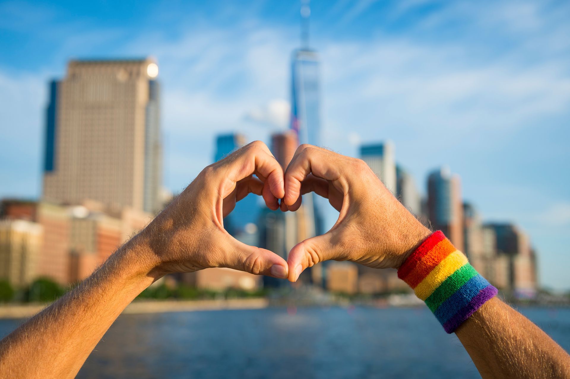 A person wearing a rainbow wristband is making a heart shape with their hands.