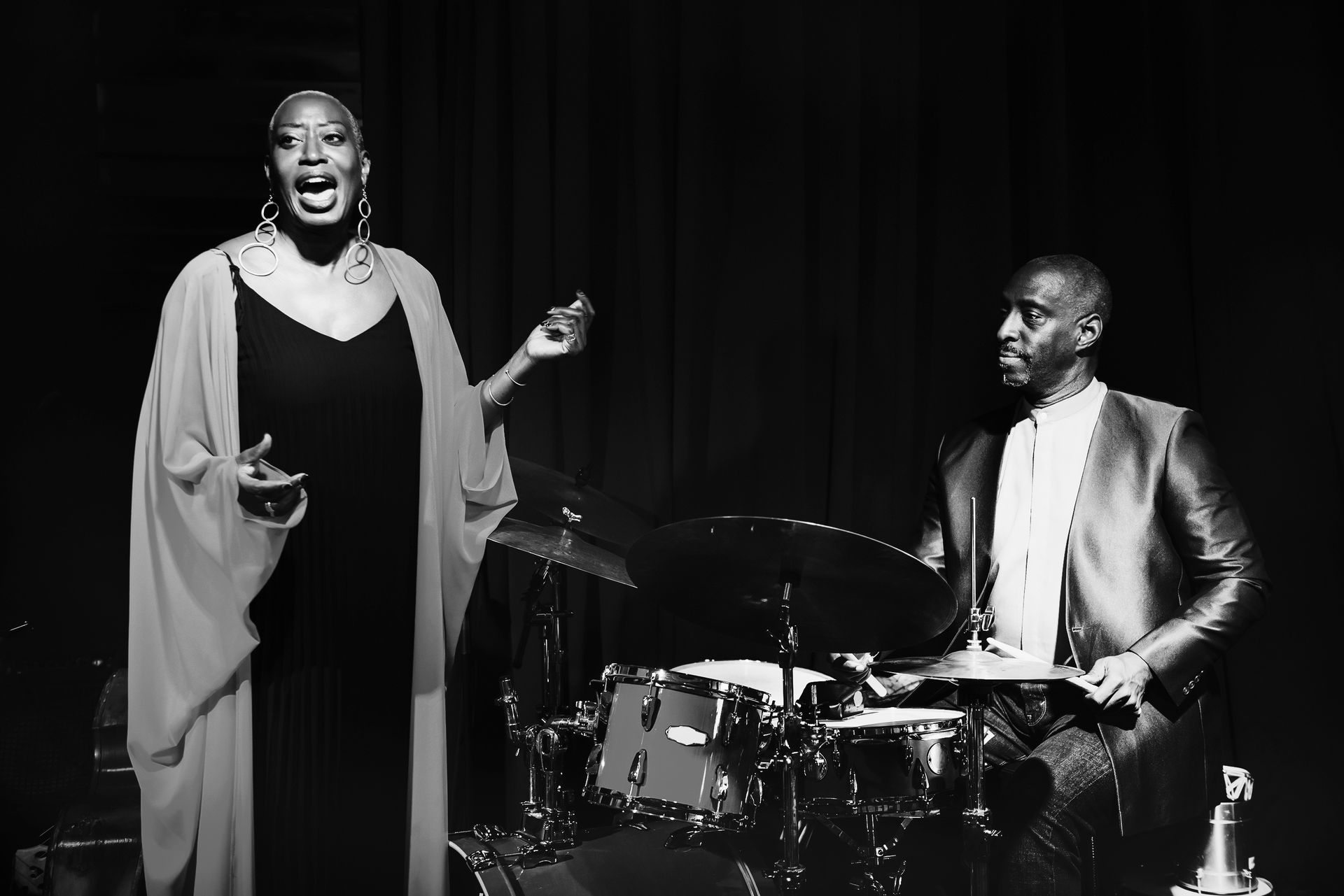 A black and white photo of a woman singing next to a man playing drums.