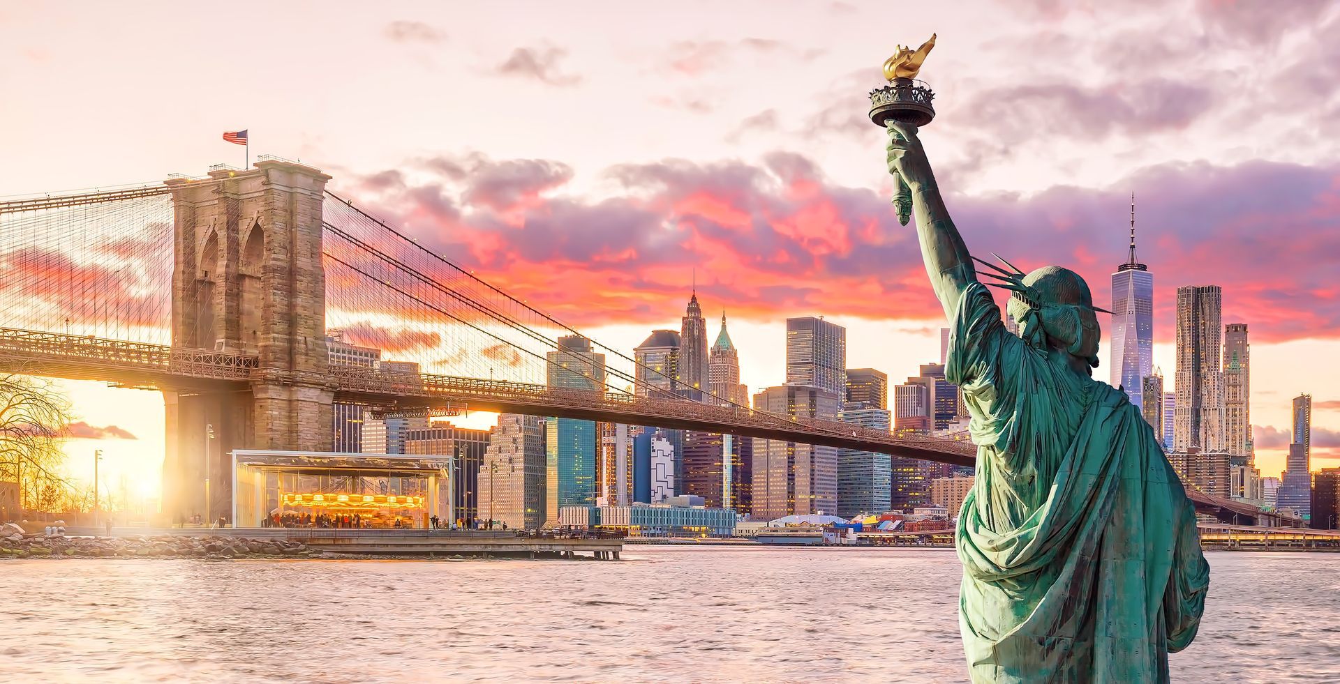Statue of Liberty with arm raised, Brooklyn Bridge, and NYC skyline at sunset.