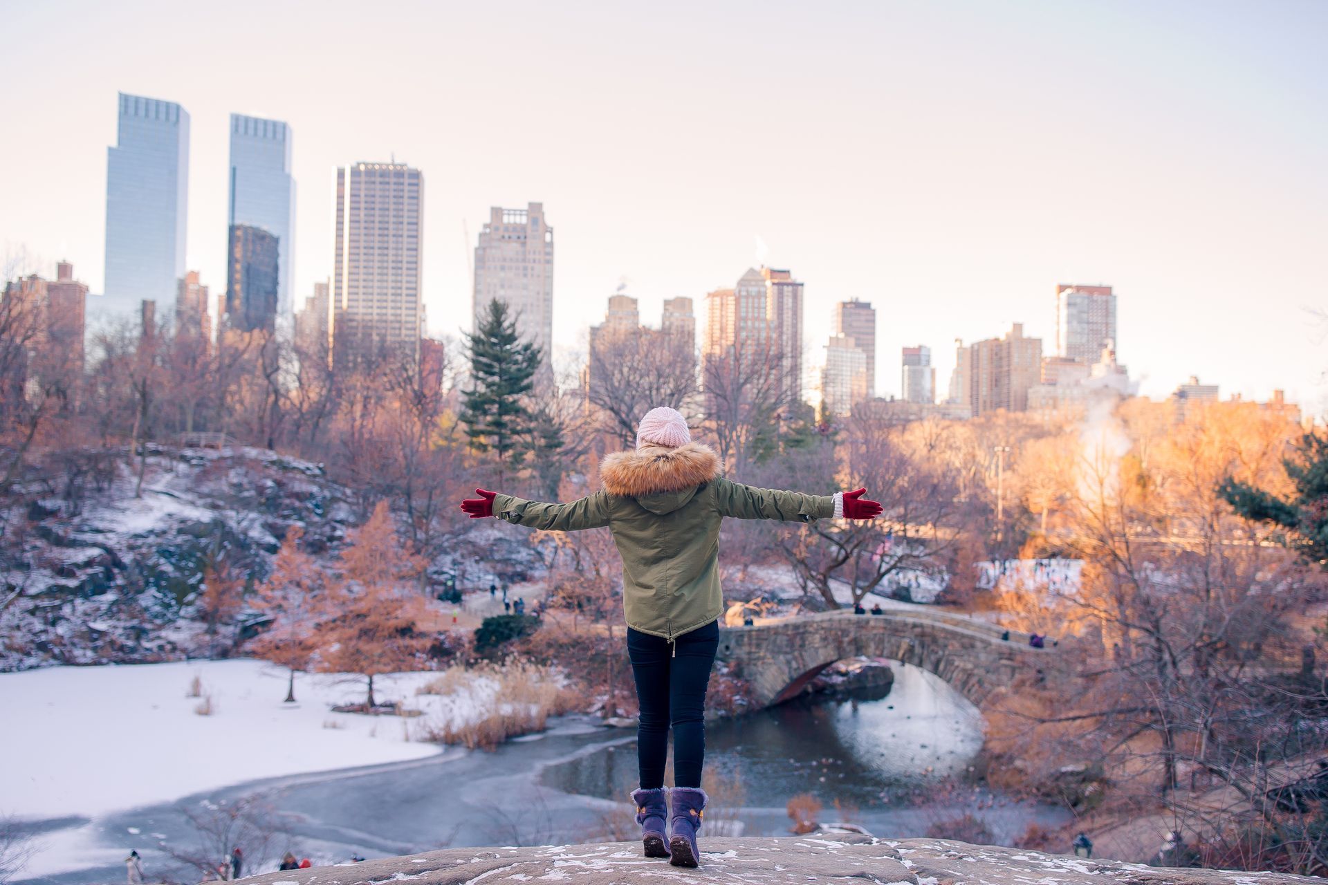 A woman is standing in a park with her arms outstretched in front of a city skyline.