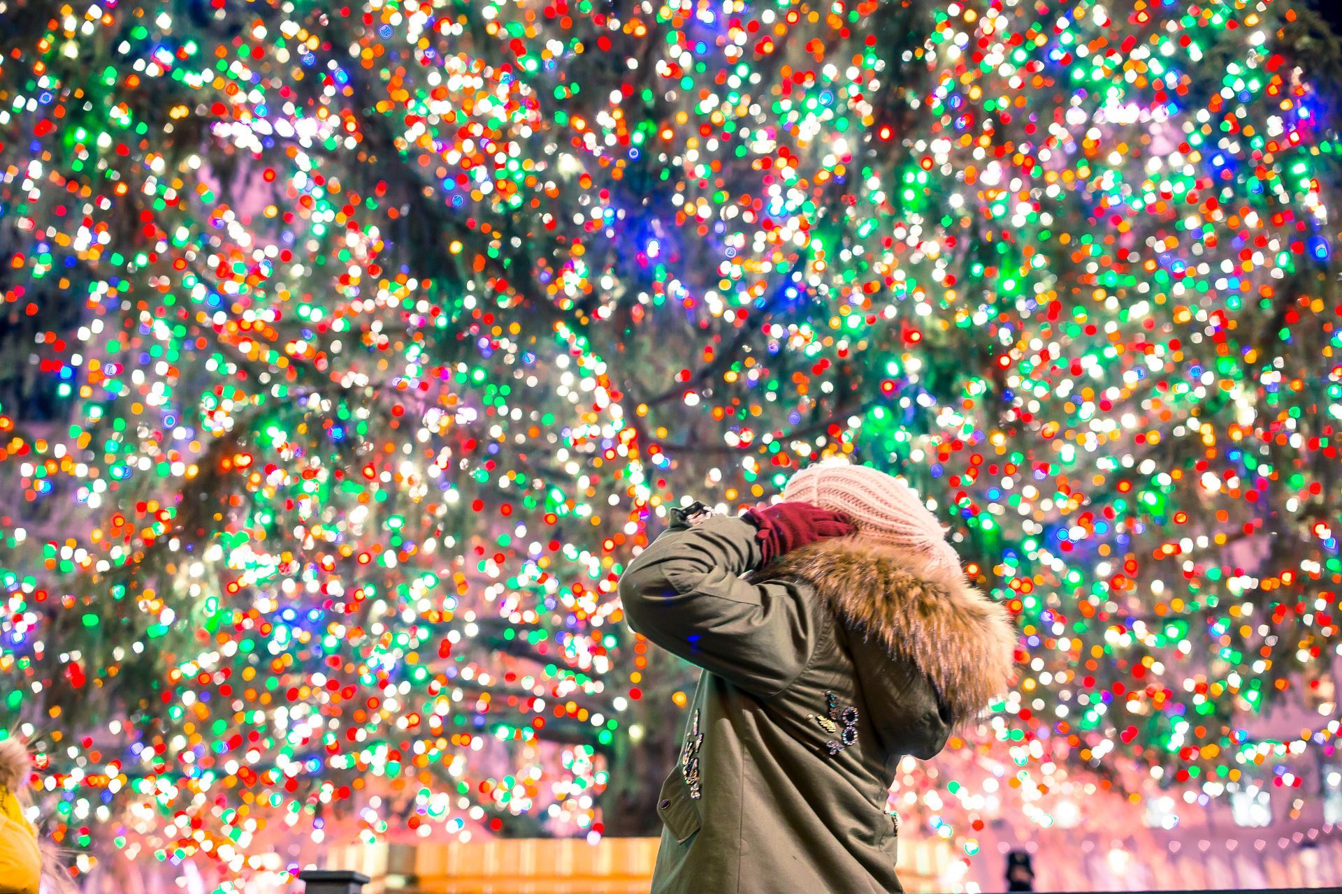 A woman is standing in front of a christmas tree with lots of lights.