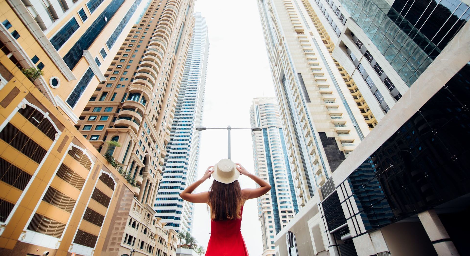 A woman in a red dress is standing in the middle of a city looking up at the skyscrapers.