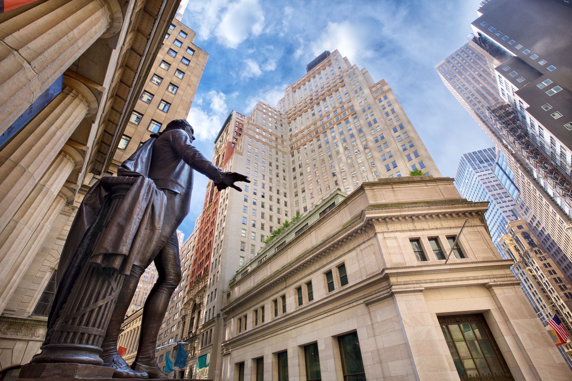 Statue of George Washington in front of tall buildings on Wall Street, New York City, under a blue sky.