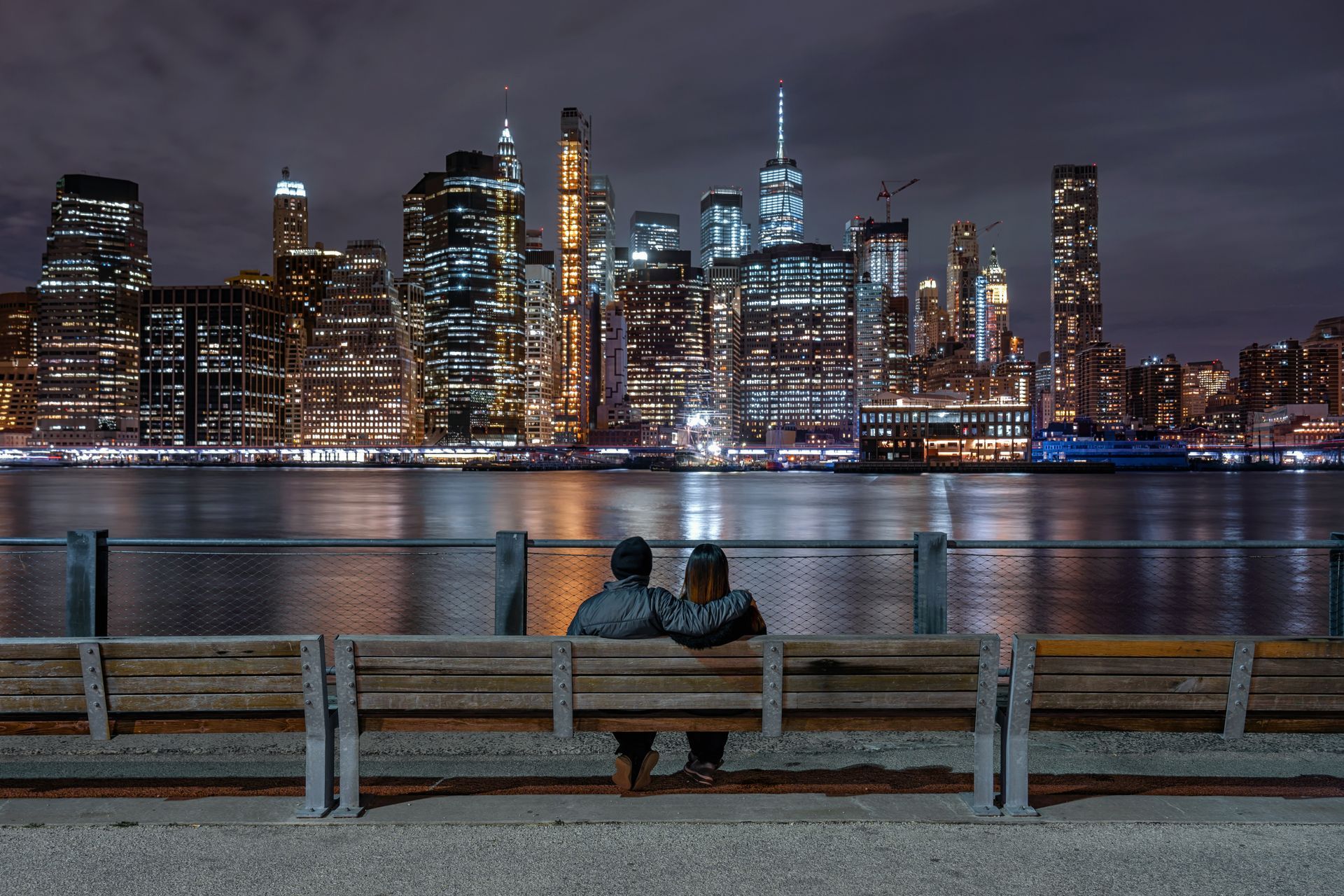 A couple is sitting on a bench looking at the city skyline at night.