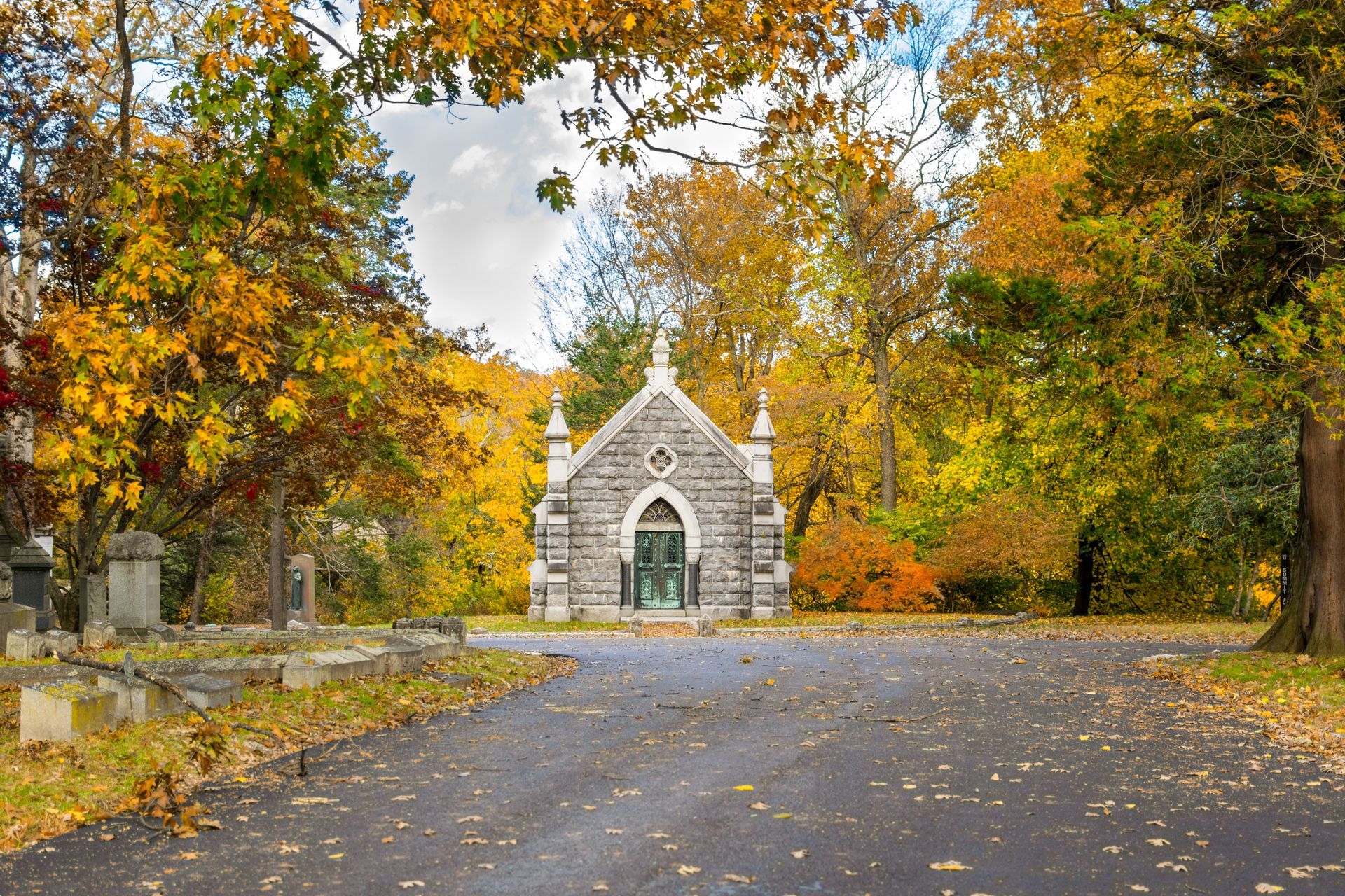 Stone chapel with green door in a cemetery during autumn. Yellow and orange foliage frame the structure.