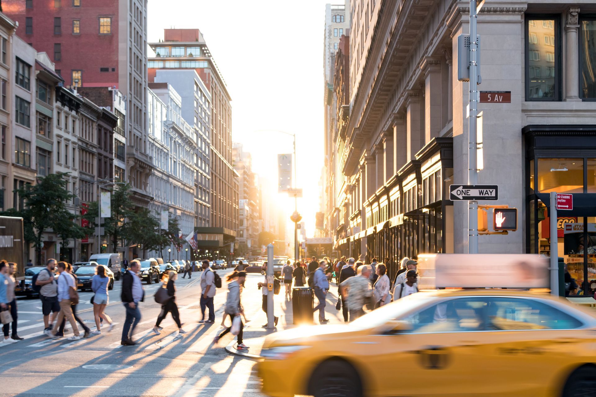 A yellow taxi is driving down a city street with people crossing the street.