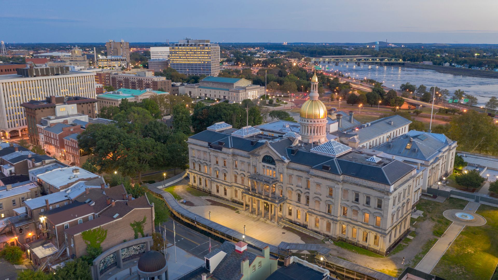Aerial view of the New Jersey State House building with a gold dome, surrounded by urban cityscape and river.