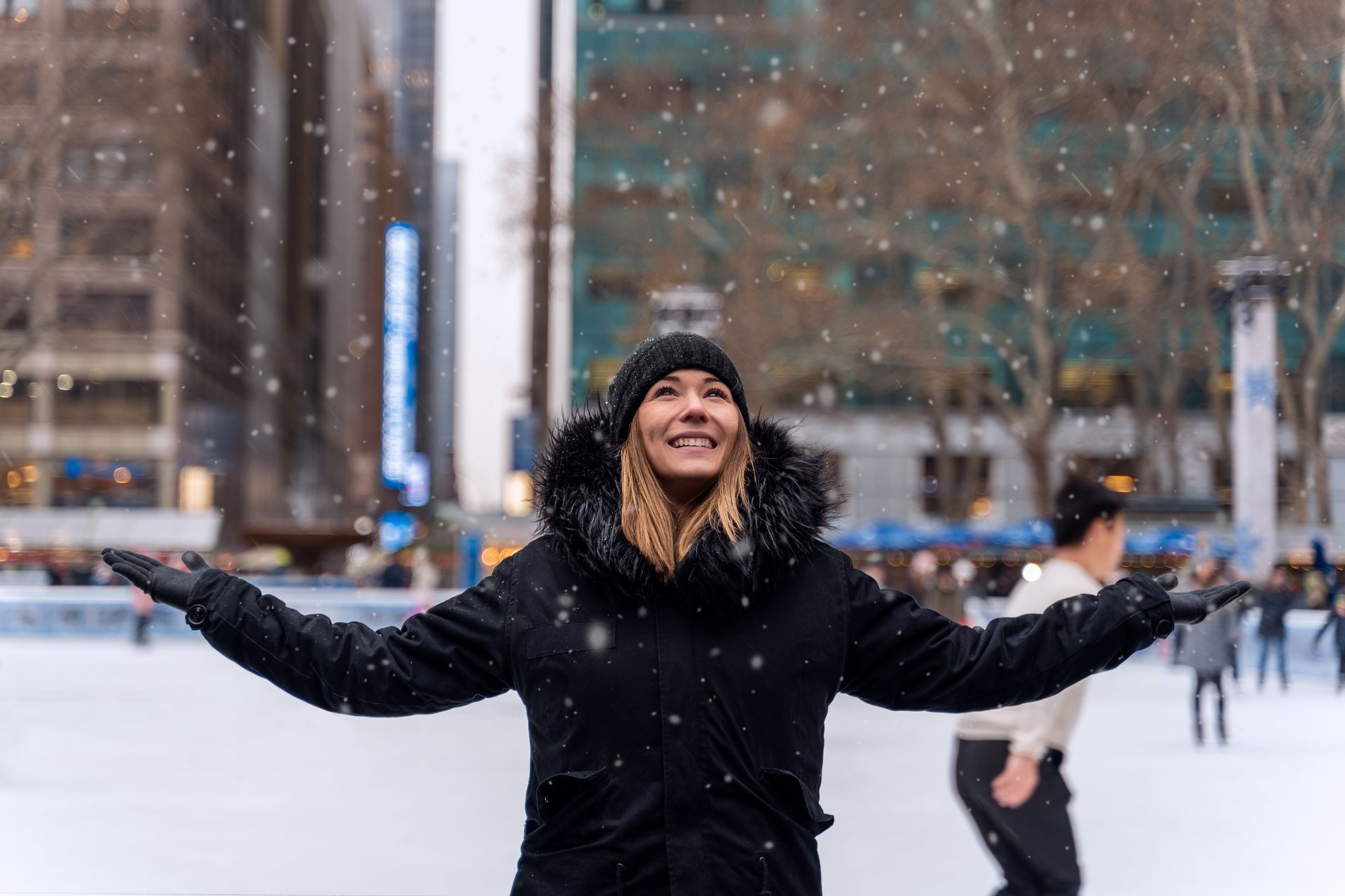 Woman with open arms, smiling, looking up at falling snow. Ice skating rink, city buildings.