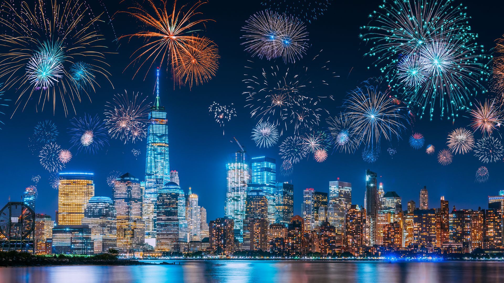 Fireworks over the illuminated New York City skyline at night, reflected in the water.