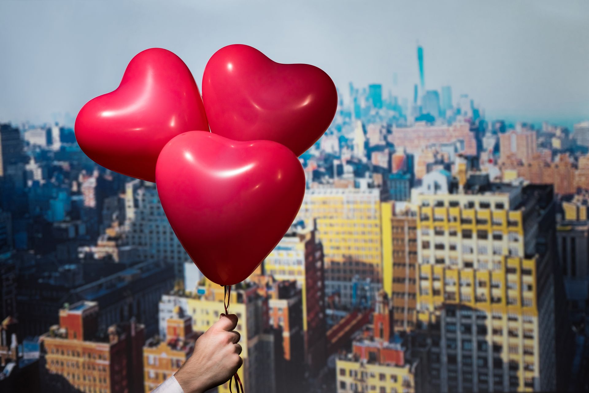Hand holding three red heart-shaped balloons, with a blurred cityscape background.