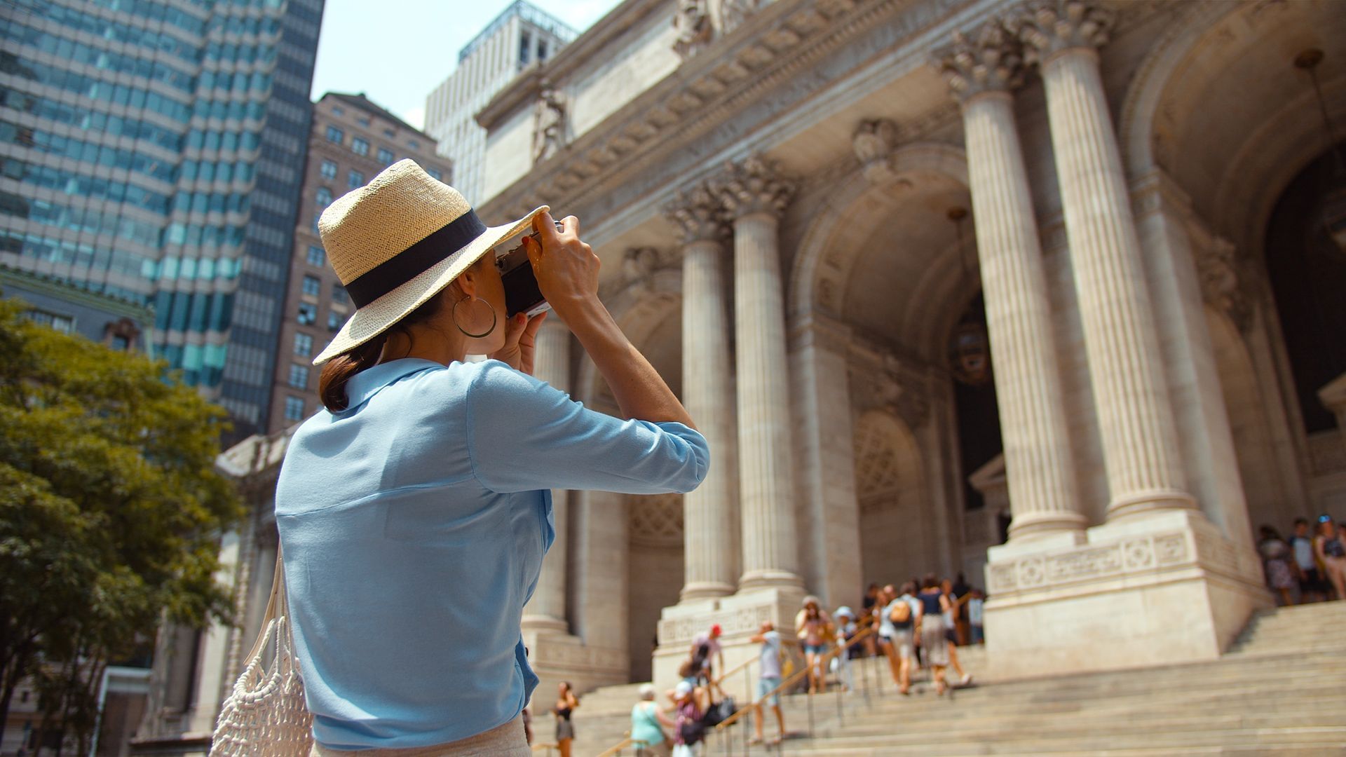 Woman in a straw hat taking a photo of a building with columns, people on steps in front.