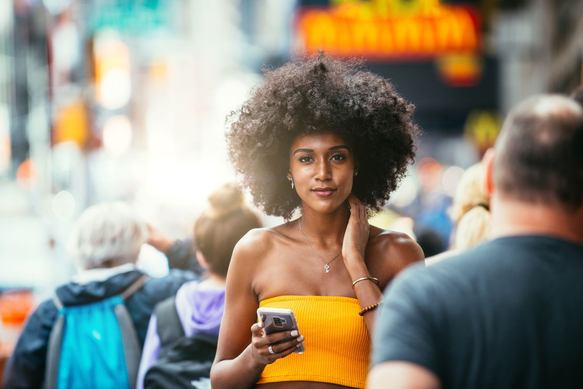 Woman with large afro in yellow top holding phone, smiles in a crowd.