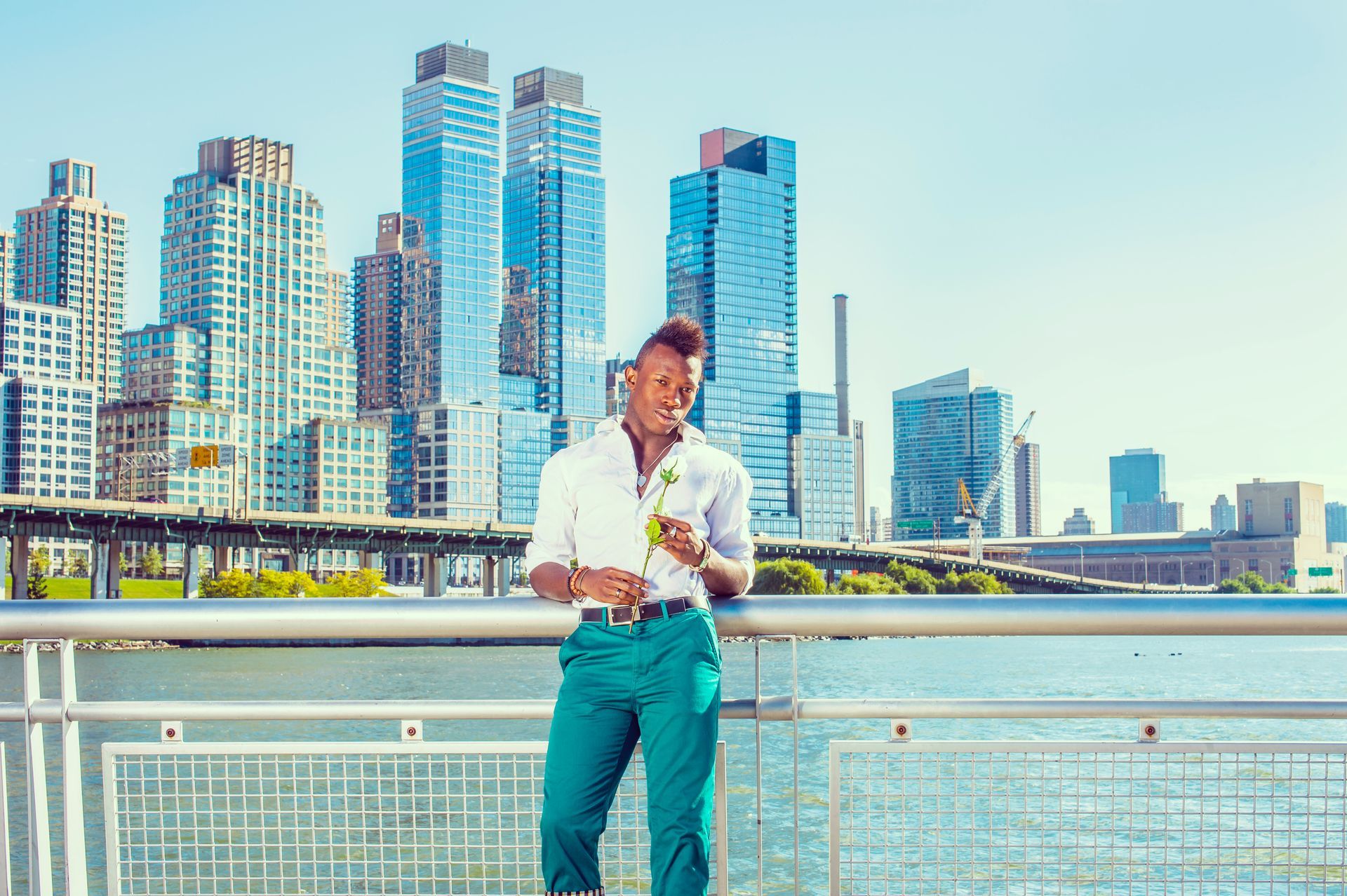 Man in white shirt and teal pants stands by a railing with city skyline and water background, holding a small plant.