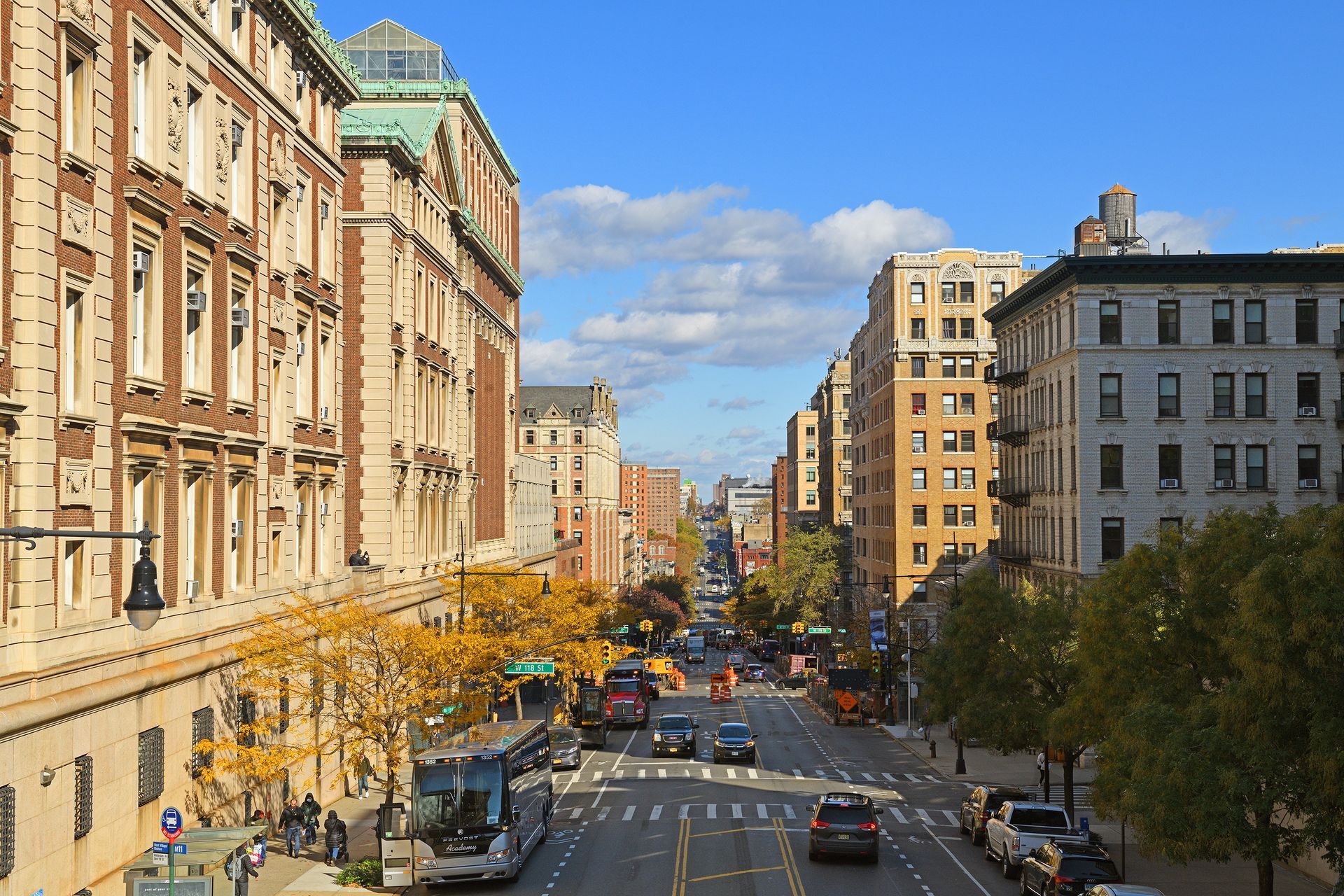 Street view with buildings, cars, and a bus under a blue sky with clouds.