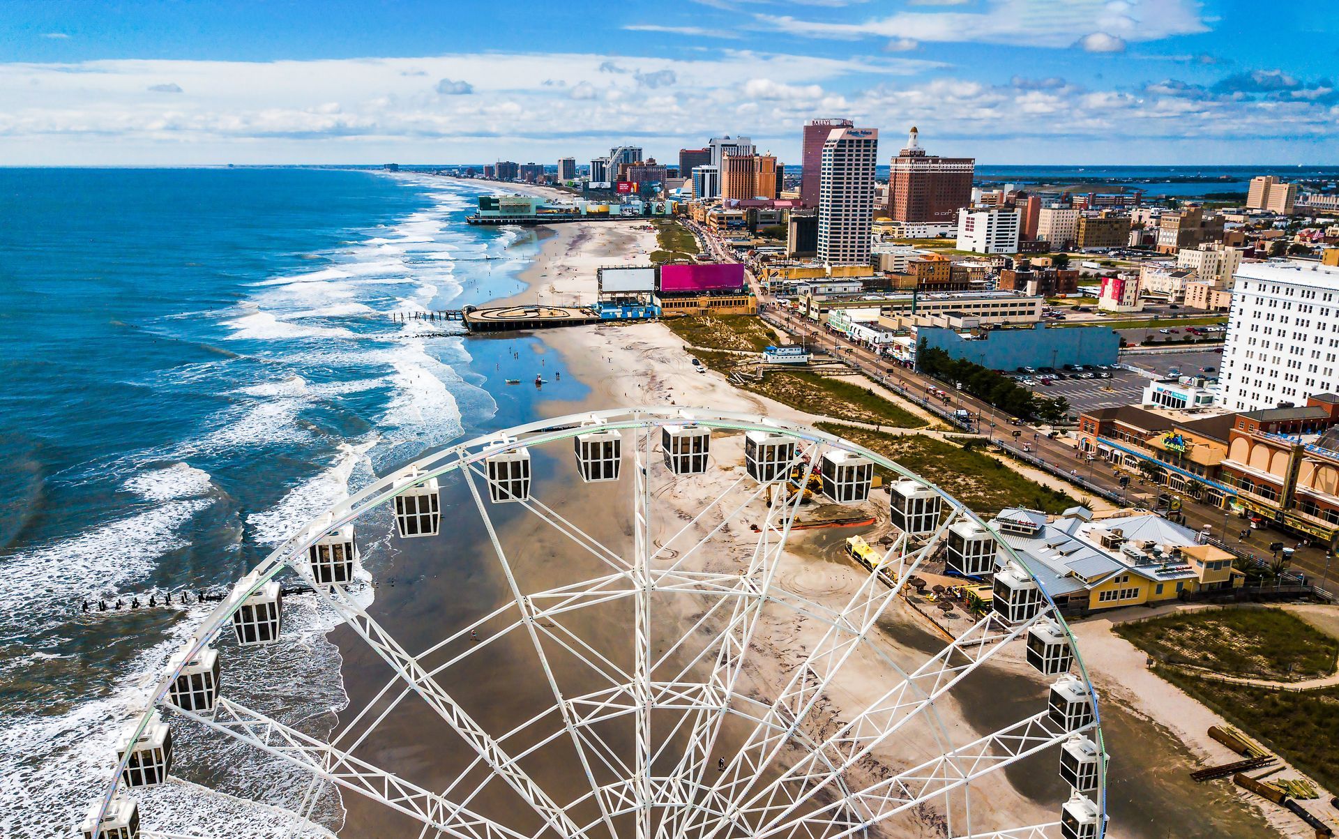 Aerial view of a beach with a Ferris wheel, boardwalk, and city buildings under a blue sky.