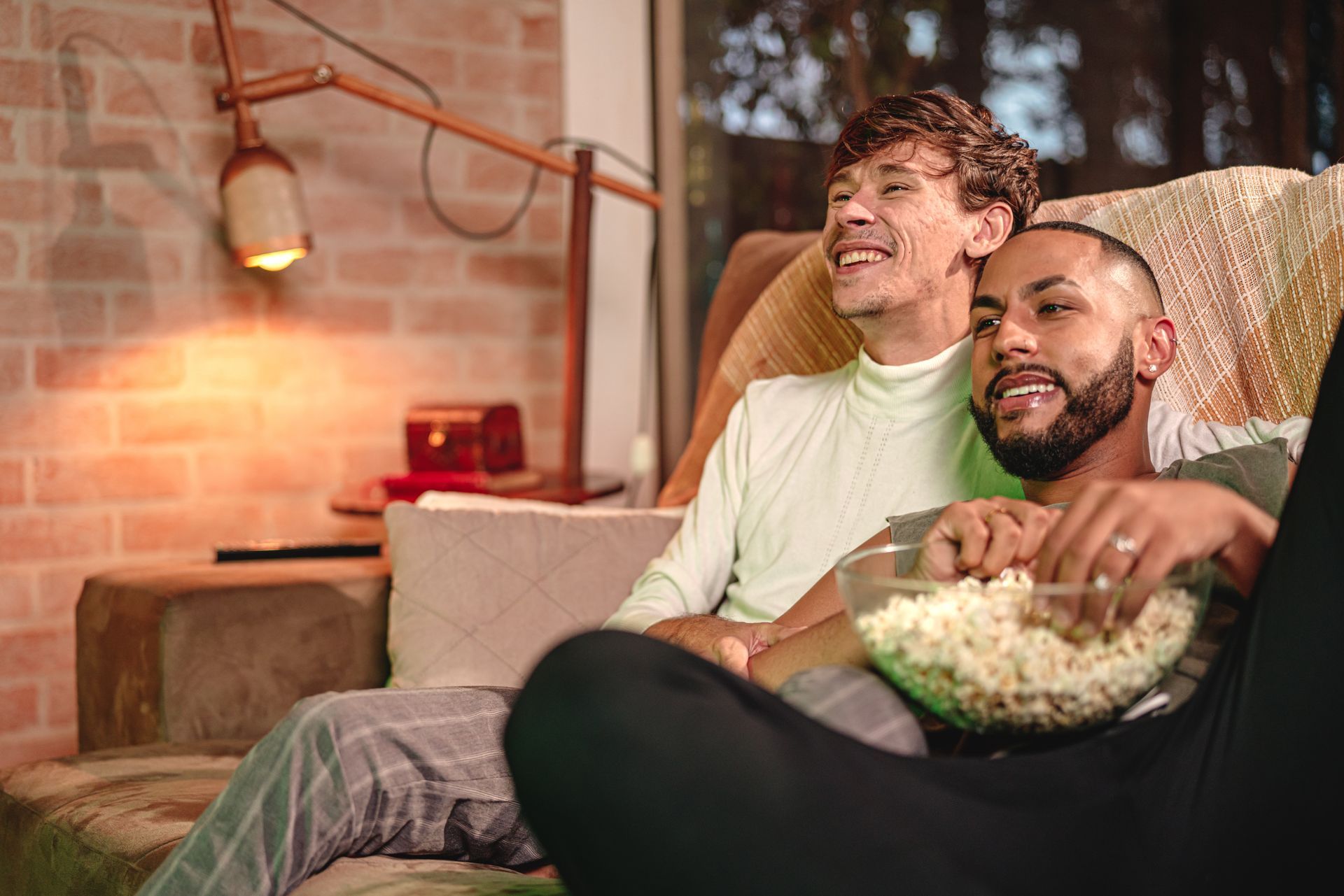 Two men are sitting on a couch eating popcorn and watching a movie.