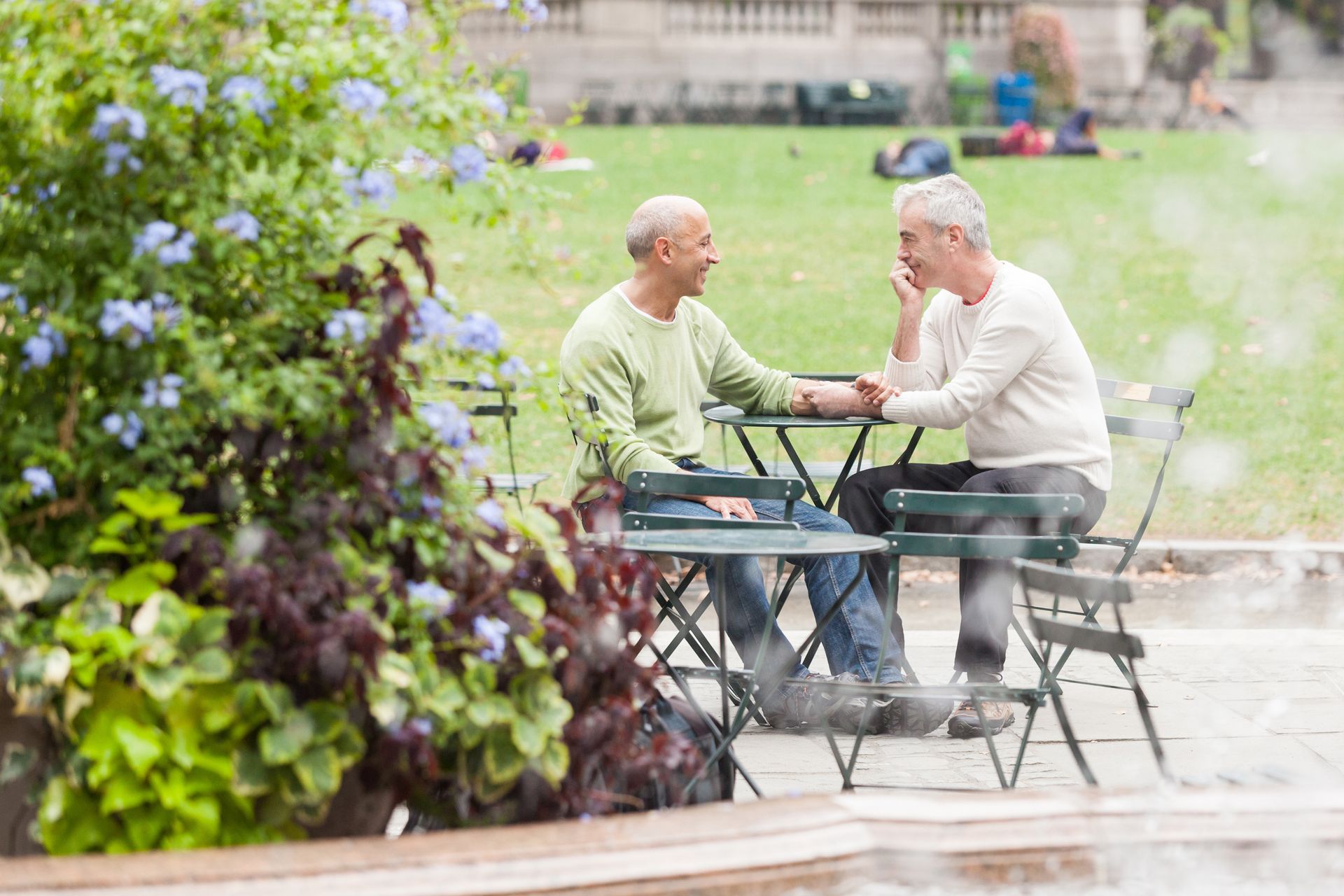 Two men are sitting at a table in a park talking to each other.