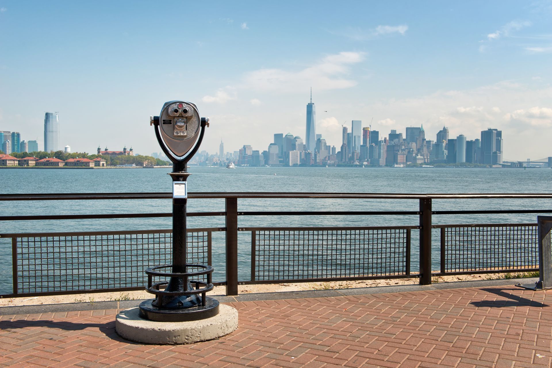 A coin-operated viewer on a brick pier overlooking the water with a NYC skyline in the distance.