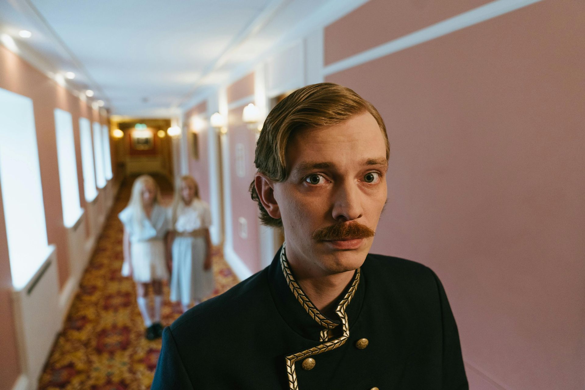 Man in hotel uniform stares, two girls in white dresses in background. Pink hallway, floral carpet.