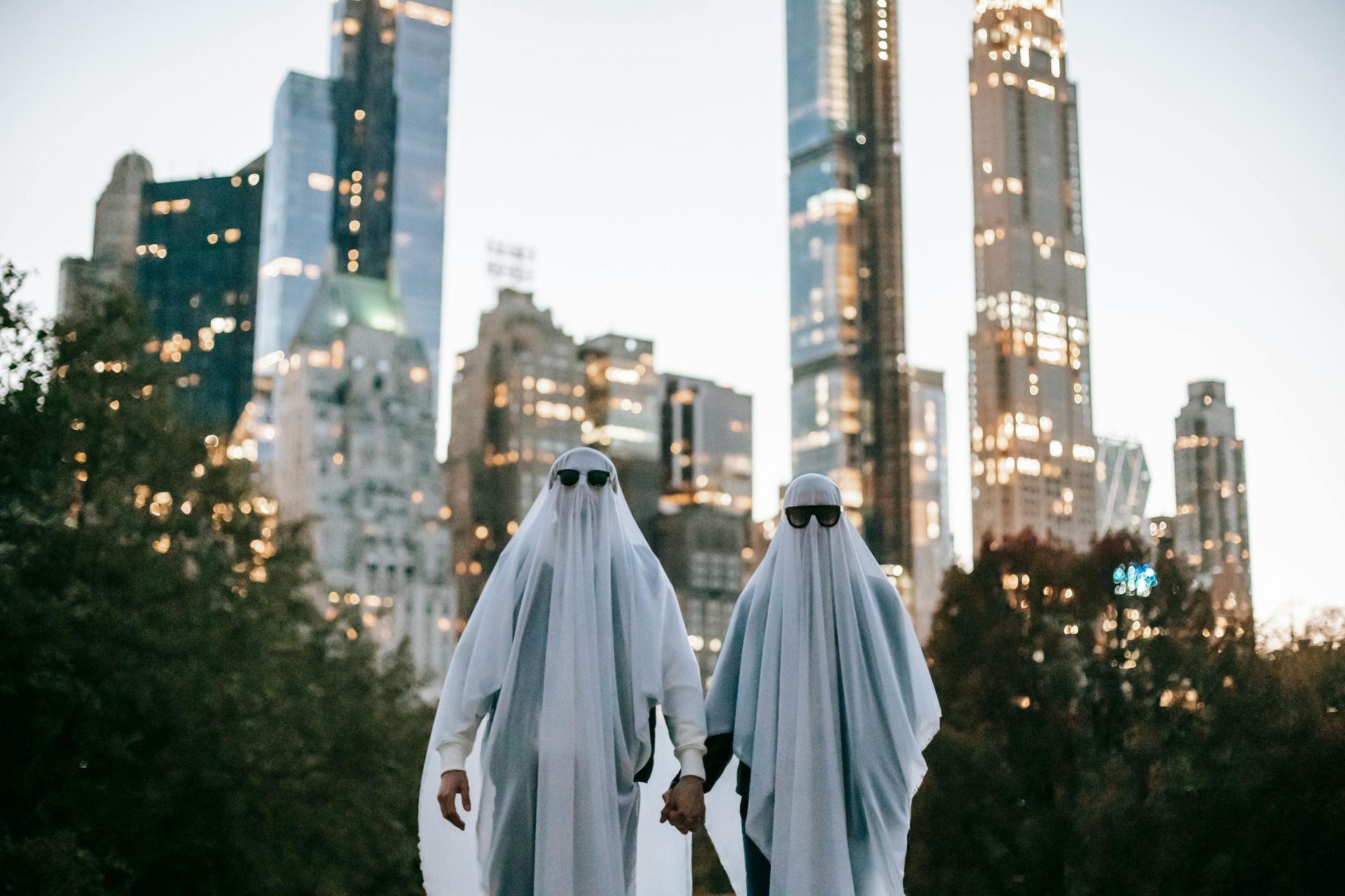 Two people in ghost costumes holding hands, wearing sunglasses, in front of city skyscrapers at dusk.