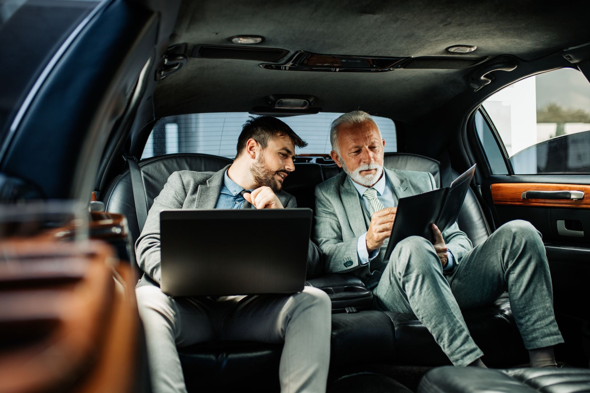Two men in suits inside a car, one using a laptop, the other a tablet, discussing work.