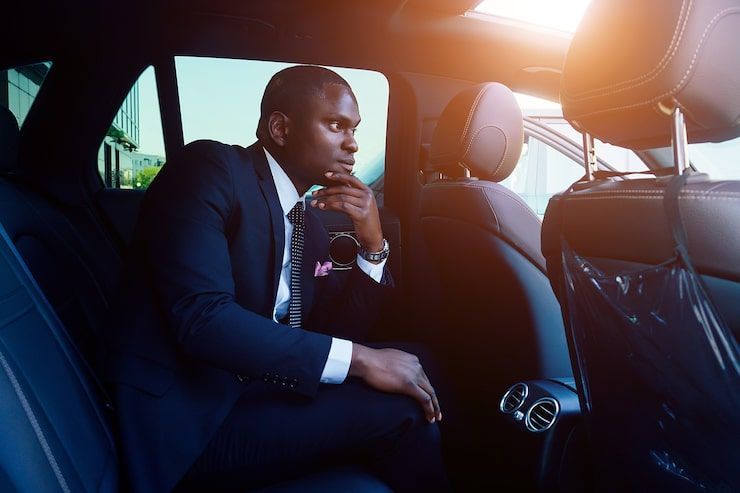 Man in a suit sits pensively in the back seat of a car, lit by sunlight.