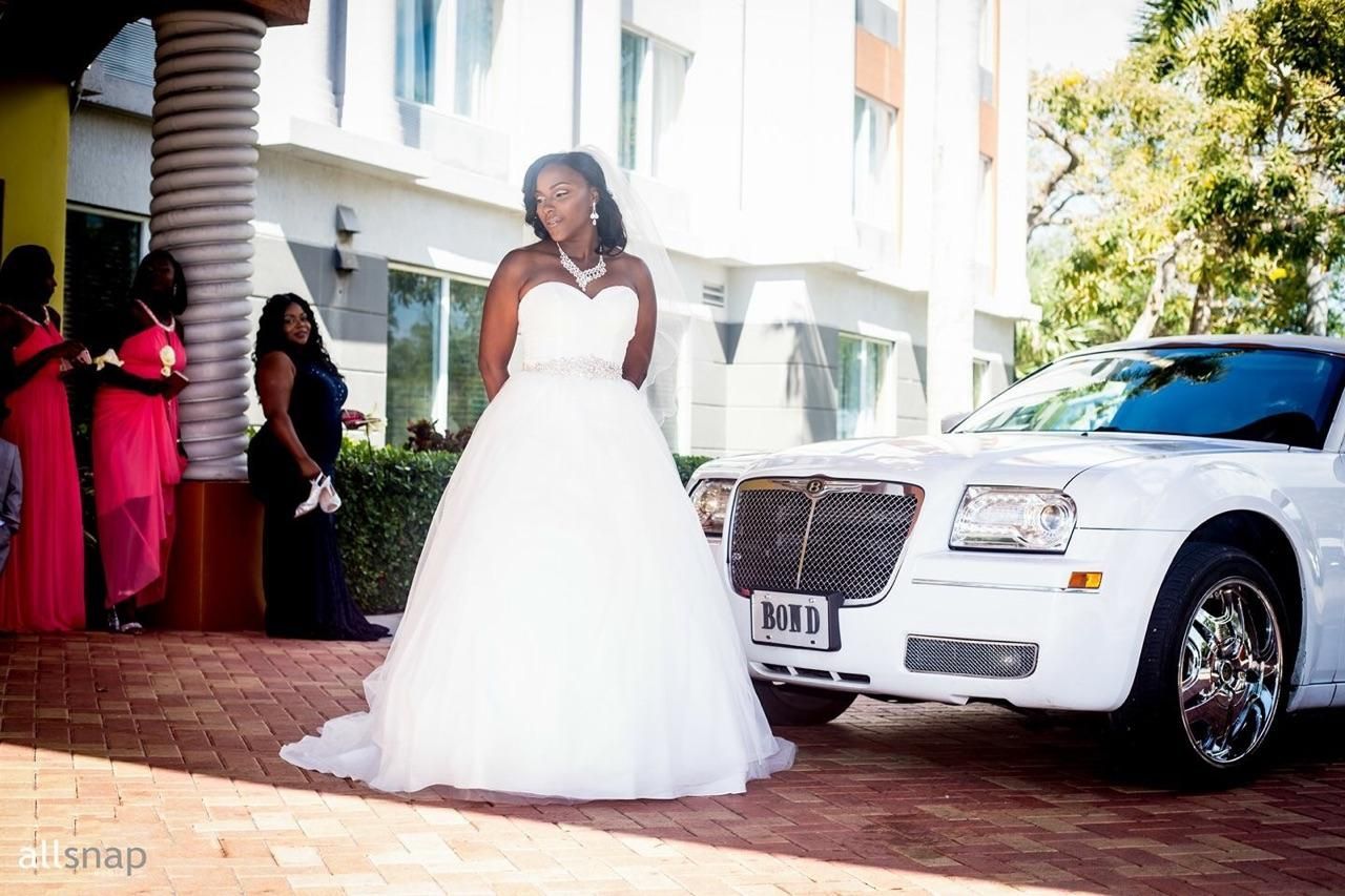 Bride in a white gown stands near a white limousine, bridesmaids stand by a building.