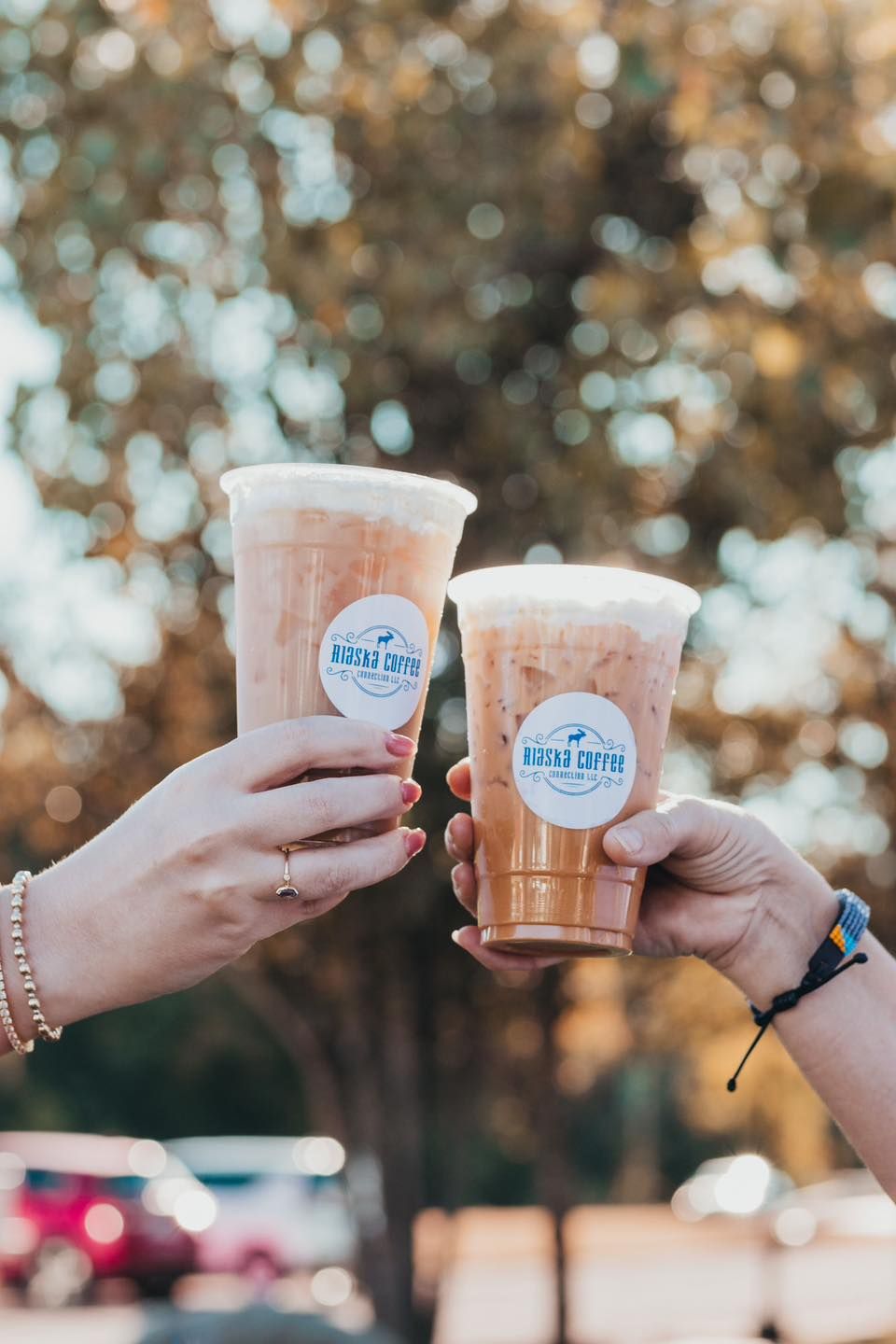 Two hands hold up iced coffee cups with branded stickers, set against a softly blurred, warm outdoor background.