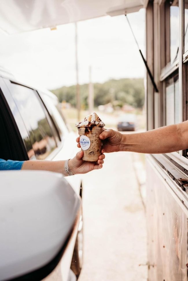 A person handing a chocolate-topped treat in a cup to a customer through a food truck window.