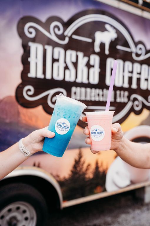 Two people holding up cups of blue and pink frozen drinks in front of an Alaska Coffee Connection food truck.