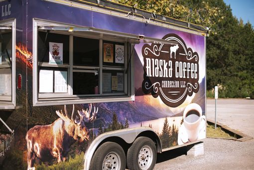 A colorful Alaska Coffee Connection food trailer featuring a moose graphic parked on a gravel lot under a bright sky.