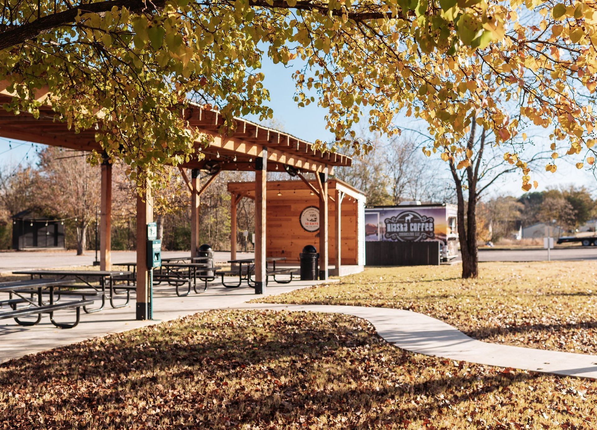 A wooden pergola covers picnic tables on a concrete patio in a park with golden autumn leaves and a food truck nearby.