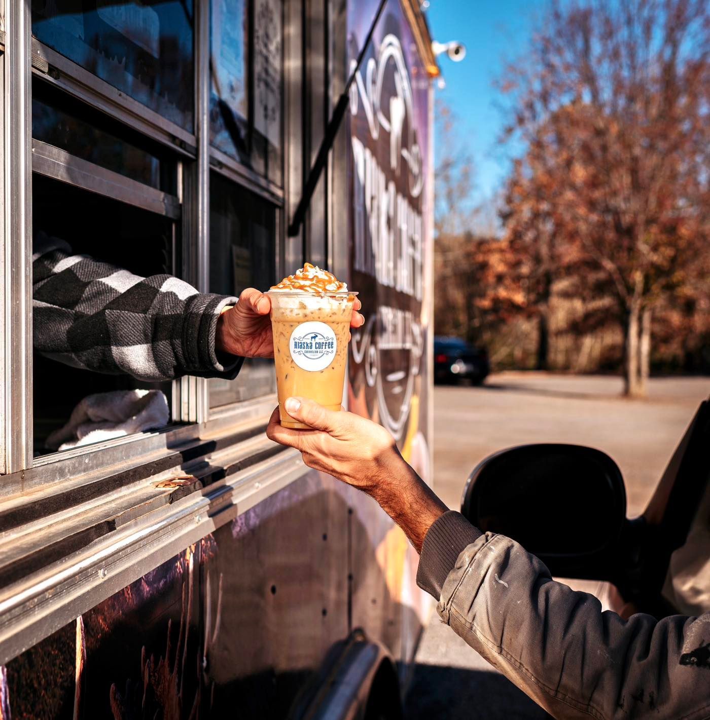 A person in a food truck hands an iced coffee drink to a customer waiting in a vehicle.